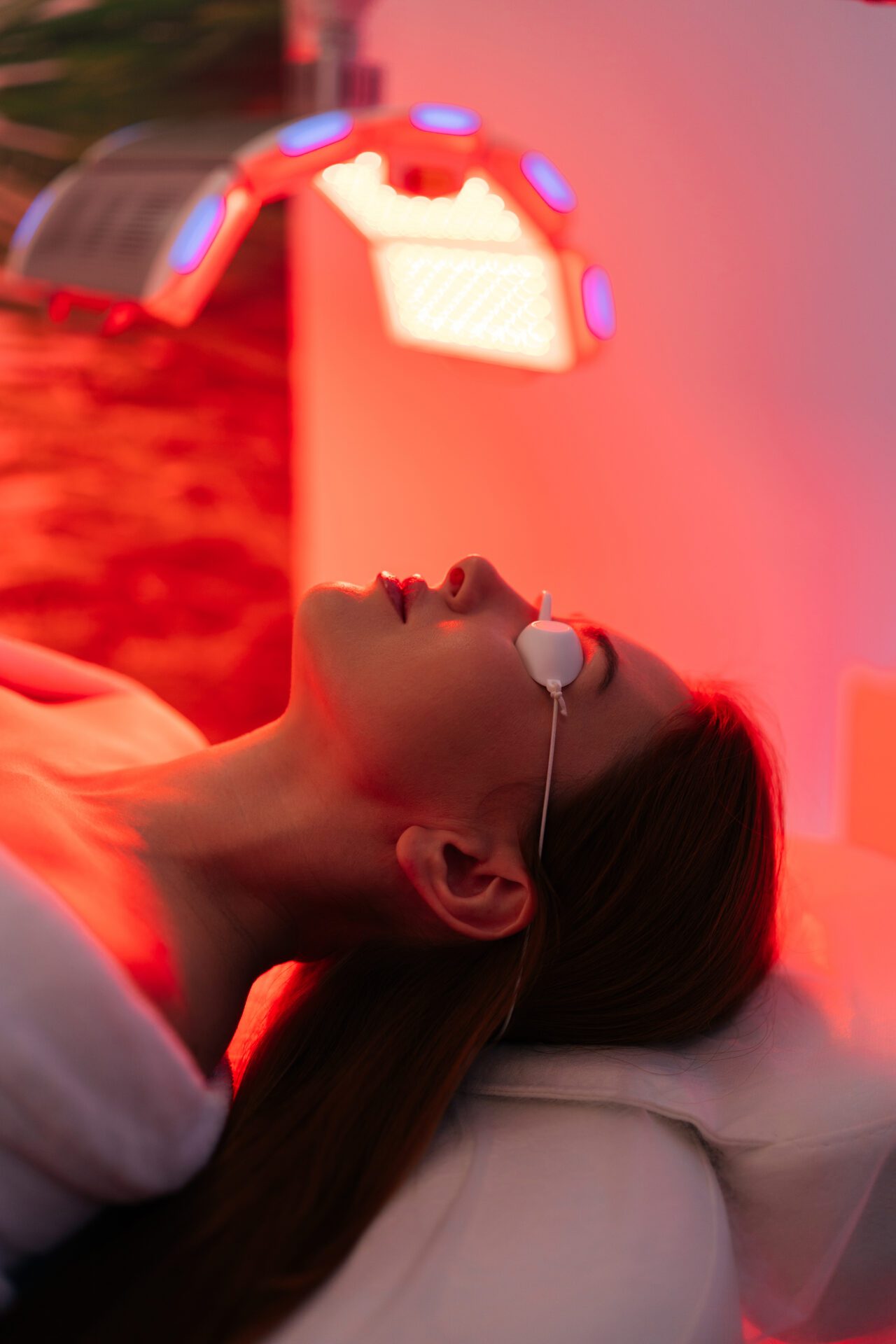 Vertical shot of young woman having red LED light facial photodynamic therapy treatment in beauty salon. Luxury medical clinic using phototherapy device You, Naturally Better